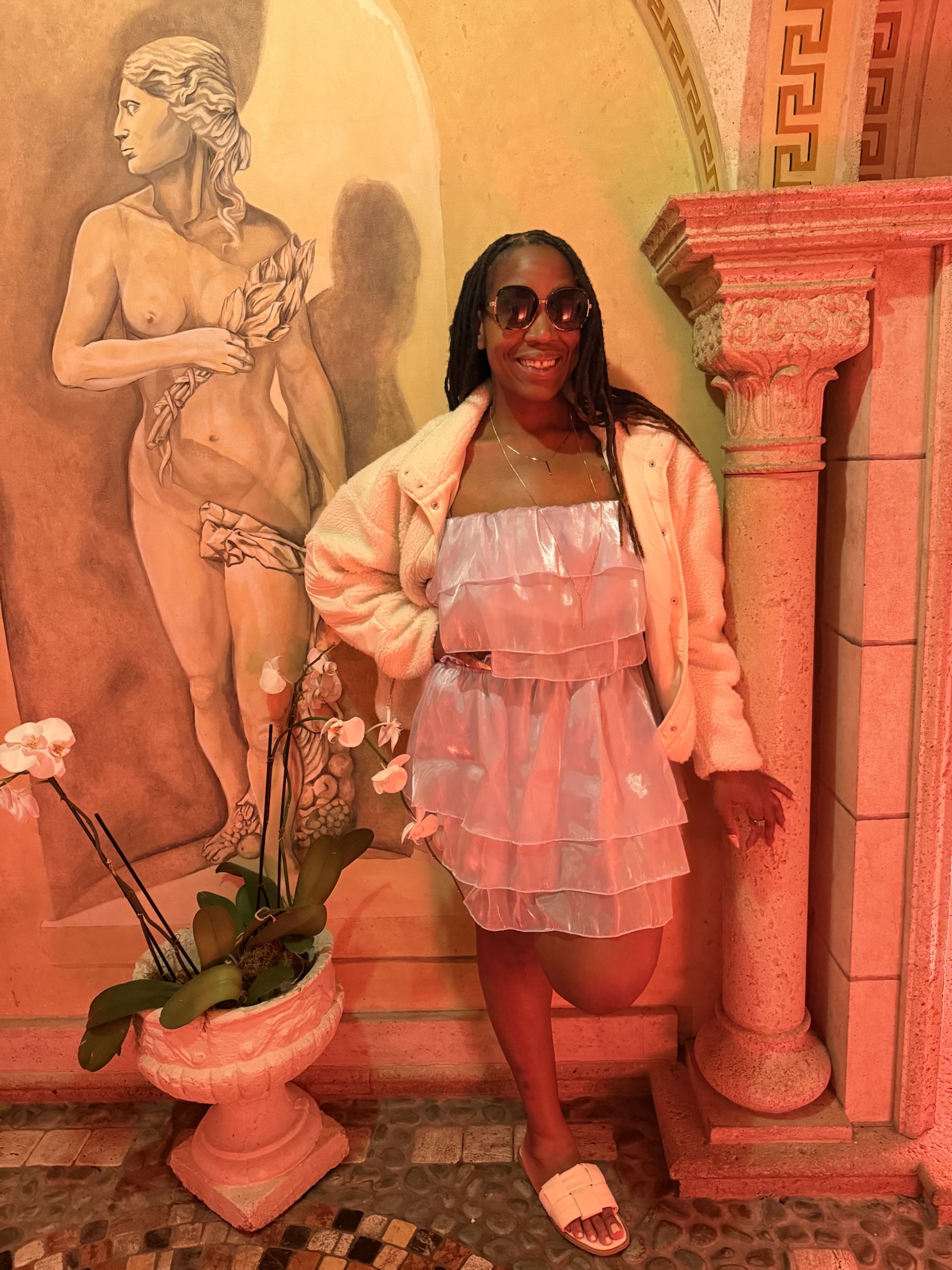 Shalaya, founder of Storm’s Mama, smiling and posing in a tiered pink organza dress and cream teddy jacket in front of a classical painted mural with orchids and a decorative column.