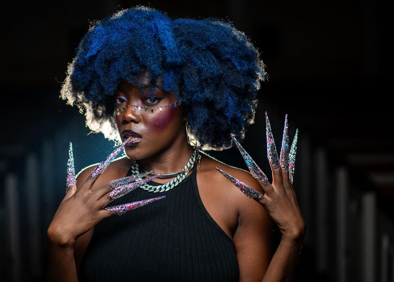 A woman with a bold blue afro and dramatic holographic stiletto nails poses confidently against a dark background, showcasing long decorated nails on both hands.