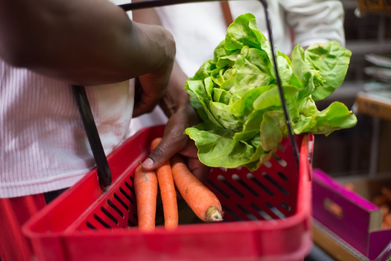 Person placing fresh lettuce and carrots into a red shopping basket at a grocery market